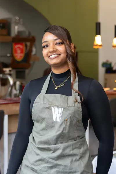Een jonge vrouw met een schort staat lachend in een horecazaak. Op de achtergrond is een bar te zien met barkrukken, koffieapparaten en verlichting.