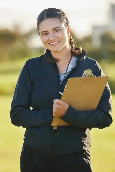 Coach staat glimlachend op een sportveld met een klembord in de hand, terwijl op de achtergrond jonge sporters in rood-witte tenues aan het trainen zijn.