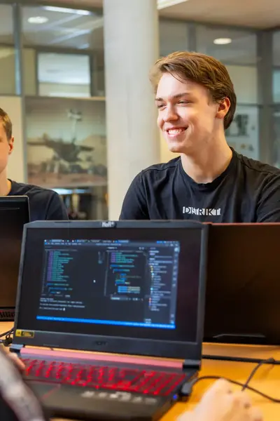 Vier studenten werken samen op laptops in een klaslokaal, met programmeercode zichtbaar op het scherm.