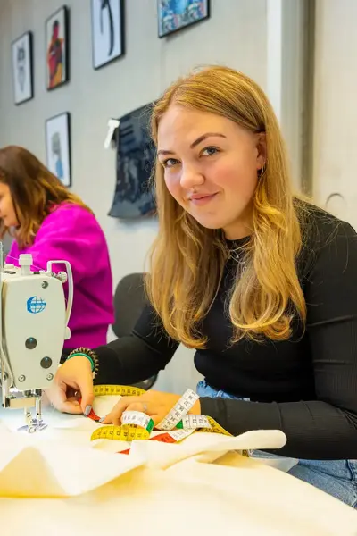 Twee vrouwen zitten achter naaimachines te werken in een hippe werkplaats of klaslokaal. De vrouw op de voorgrond lacht in de camera terwijl ze bezig is.