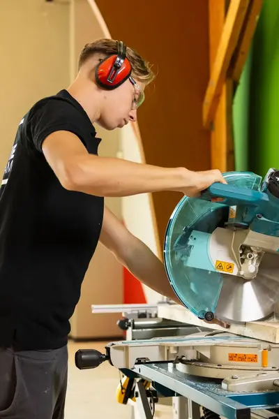 Student met oorbescherming op staat in een praktijklokaal en is aan het werk met een zaagmachine.