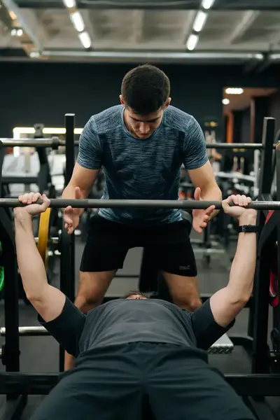 Man is in sportschool bezig met bankdrukken. Een trainer helpt hem daarbij.