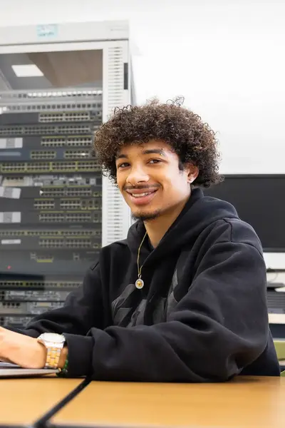 Student werkt op een laptop in een ICT-lokaal met netwerkapparatuur op de achtergrond.