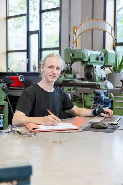 Student met laptop en schrift in werkplaats.